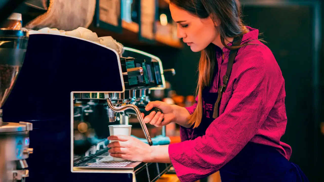A female barista brewing coffee wearing a CoffeeOwl branded apron with a central logo.