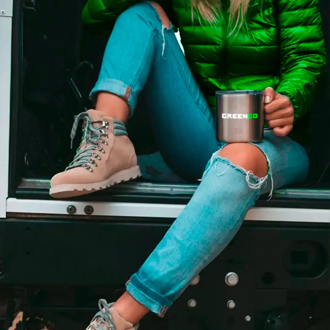 A woman in a green outdoors jacket sitting in a car trunk and holding a GreenGo branded steel mug.