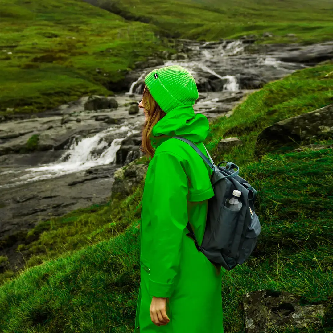 A woman, wearing a green outdoors jacket, a winter hat and a gray backpack, standing in front of a beautiful river in mountains.