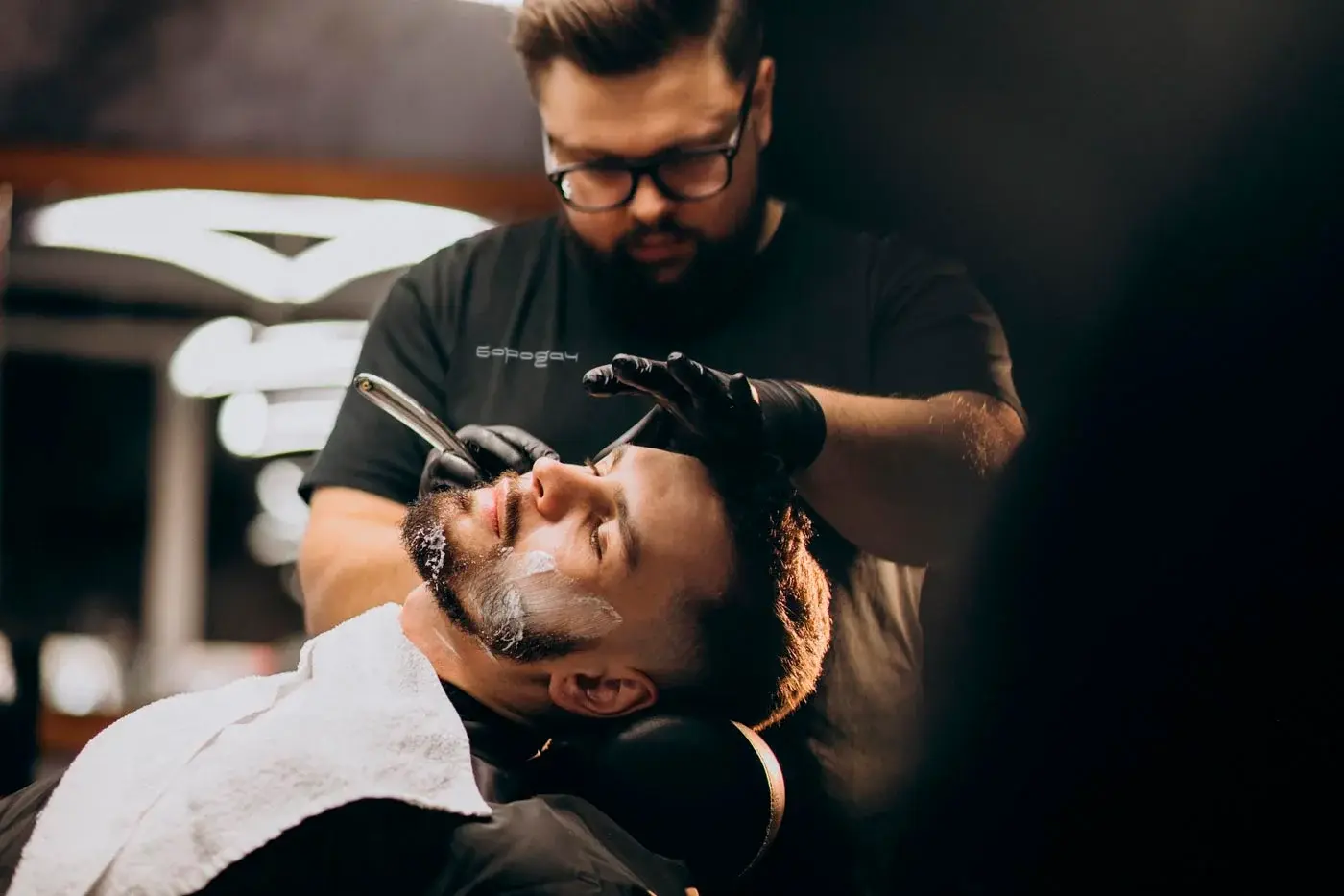 A barber in a Borodach-branded shirt shaving a client's face.